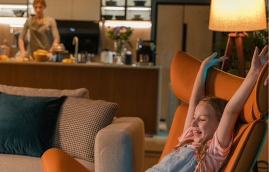 Young girl laughing on orange arm chair while mum cooks in kitchen in background